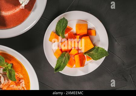 Zucca tagliata a dadini con menta su un piatto bianco con zuppa di verdure, purè di patate e pancake su fondo testurizzato grigio. Foto di alta qualità Foto Stock