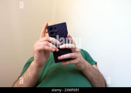 Uomo che usa un nuovo telefono cellulare, tiene il dispositivo davanti al viso, indossa una polo verde. Persona non identificata vista dal basso, sfondo bianco Foto Stock