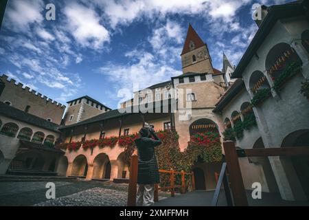 Austria - 26 ottobre 2023: Una donna con un cappotto nero si erge su una ringhiera di legno di fronte ad un edificio storico, con un cielo blu e nuvole soffici behi Foto Stock