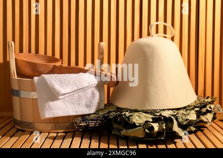 Frusta per sauna in quercia, cappello in lana di feltro, secchio, asciugamano e mestolo su una superficie in legno Foto Stock