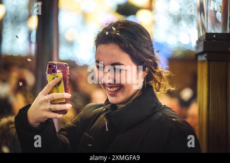 Una giovane donna gioiosa con il cappotto nero scatta selfie con lo smartphone in un ambiente vivace al chiuso, un evento festivo. Che riflette la felicità, il freddo, e. Foto Stock
