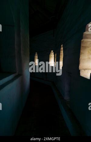 Uno stretto corridoio in un edificio storico, poco illuminato da piccole finestre lungo il muro, che creano un ambiente misterioso e antico. Foto Stock