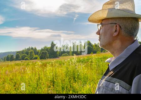 Un uomo che indossa un costume da apicoltura e un cappello di paglia si siede sull'erba in un campo illuminato dal sole, godendosi una pausa. Il paesaggio sereno e la luce del sole si illuminano Foto Stock