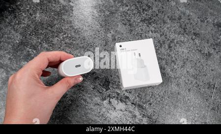 Jurmala, Lettonia 22.06.2024: Donna con un caricabatterie Apple di tipo c. scatola bianca dell'adattatore di ricarica originale Apple da 20 W su un tavolo grigio chiaro. Foto Stock