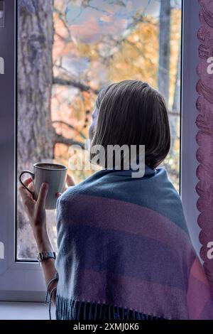 Donna in scialle viola caffè alla finestra, contemplando e contemplando il paesaggio autunnale. vista posteriore. Foto Stock