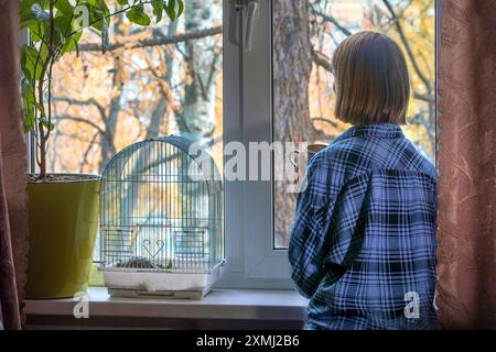 Donna in camicia blu a quadri che beve un caffè alla finestra con un paesaggio di caduta coperto. vista posteriore. Foto Stock