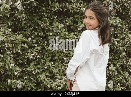 Una ragazza dai capelli lisci con una camicia bianca si posa con la schiena alla telecamera e la testa girata, sorridente, in un giardino lussureggiante. Foto Stock