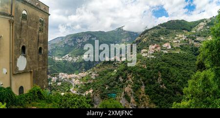 Una vista panoramica di un villaggio annidato sulla Costiera Amalfitana con lussureggianti colline verdi e nuvole sparse nel cielo. Foto Stock