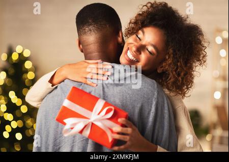 Grati afro donna con un regalo di Natale abbracciando il marito a casa Foto Stock