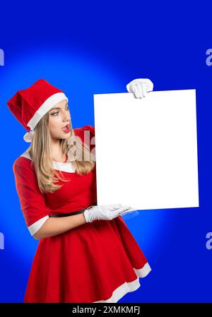 Giovane donna bionda vestita da Babbo Natale con un cartello sorpreso su sfondo blu Foto Stock