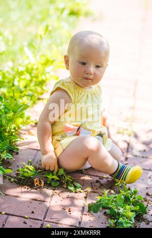 Una bambina in abito giallo siede su un sentiero in mattoni, circondata da piante verdi e luce solare brillante. Foto Stock