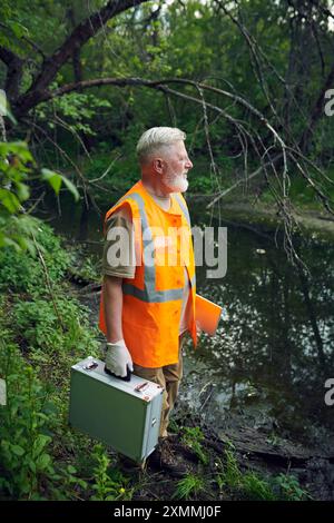 Vista verticale ad angolo alto alto, colpo lungo dell'ecologista senior con kit da laboratorio portatile in piedi sulla riva del fiume nella foresta Foto Stock