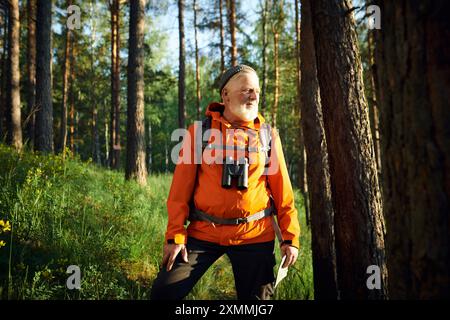 Ritratto medio lungo, alto turista caucasico, con giacca arancione e berretto in piedi nella foresta di montagna che guarda lontano Foto Stock