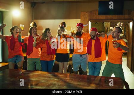 Amici, tutti in camicie arancioni e sciarpe rosse, bicchieri allegri, canto di gioia e felicità, celebrando la vittoria della squadra preferita al bar. Foto Stock