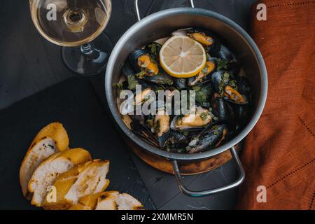 Cozze al vino con prezzemolo e limone tritati, un bicchiere di vino bianco e una baguette tostata su un tavolo grigio. Piatti tradizionali francesi e italiani. Ad alta velocità Foto Stock