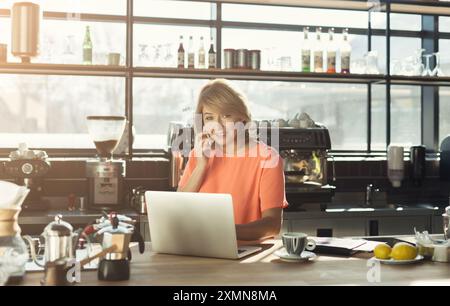 Barista di mezza età che lavora con un laptop e parla con uno smartphone Foto Stock