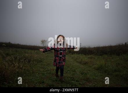 Bambina felice che corre su un campo nebbioso con un cappotto a quadri Foto Stock