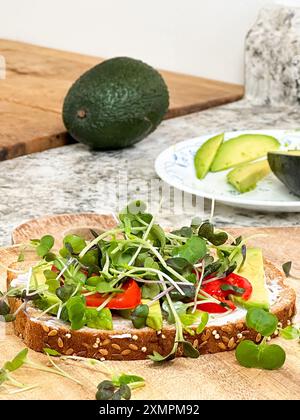 Viene preparato un nutriente panino a faccia aperta con una base di pane integrale con semi di sesamo, ricoperto di avocado a fette, verdure fresche e pomodoro ciliegino Foto Stock