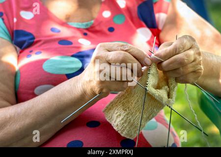 Mani di una donna anziana che lavora a maglia calze per nipoti. Pensionessa che si diverte il suo hobby in cortile durante il sole del giorno d'estate. Primo piano. Foto Stock