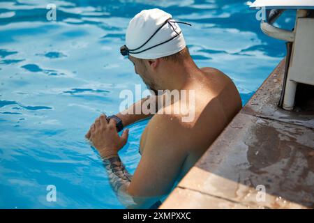 La foto dell'uomo nella vista posteriore con il berretto bianco e gli occhiali di protezione esamina il suo smartwatch mentre si riposa sul bordo della piscina, circondato da acque cristalline. Foto Stock