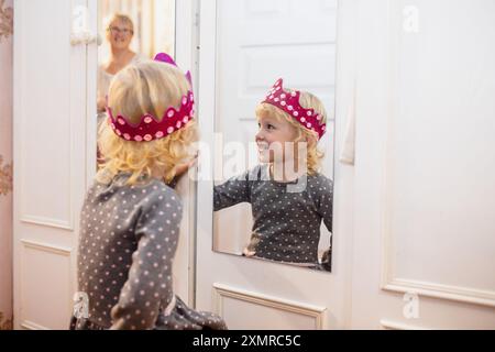 Una giovane ragazza allegra con un abito punteggiato di polka ammira il suo riflesso mentre indossa una corona rosa brillante. La sua espressione gioiosa riflette l'eccitazione Foto Stock