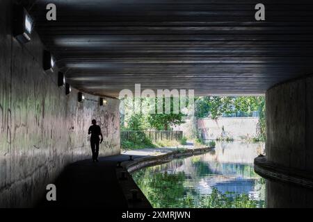 Vista di una donna solitaria che cammina accanto a un canale del Regno Unito usando il sottopassaggio/tunnel buio, durante il giorno. Foto Stock