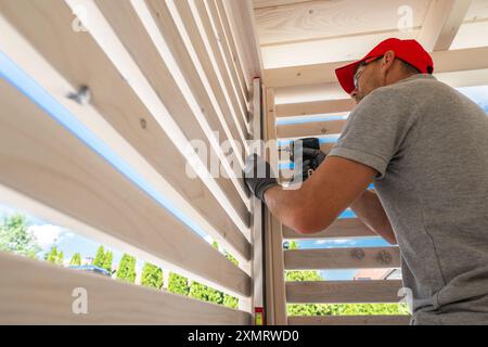 Un lavoratore esperto si concentra sull'installazione di spranghe in legno in un gazebo sul retro. Il sole splende brillantemente, illuminando lo spazio di lavoro come l'artigiano usa Foto Stock