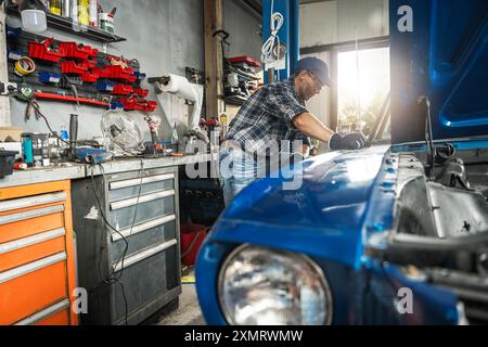 Un meccanico esperto si concentra sulla riparazione di una classica auto blu in un'officina ben organizzata. Gli attrezzi e le attrezzature lo circondano durante la valutazione del Foto Stock