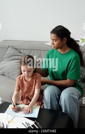 Una ragazza afroamericana in un abbigliamento casual seduto sul divano accanto a un'adorabile bambina che disegna un'immagine del suo animale domestico con pastelli Foto Stock