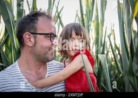 primo piano ritratto di un padre che tiene una figlia in abito rosso in piedi nel campo di mais Foto Stock