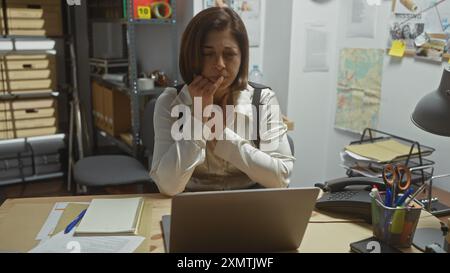 Detective ispanica pensiva in ufficio con portatile, tabellone delle prove e fascicoli. Foto Stock