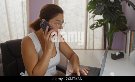 Donna che parla al telefono mentre utilizza il computer portatile in un ufficio domestico, che mostra un'espressione mirata e capelli bruna, seduta sulla sedia dell'ufficio con la penna Foto Stock