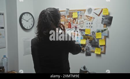 Una giovane donna indaga su un crimine in una stazione di polizia, analizzando le prove su un tabellone mentre è al telefono, al chiuso. Foto Stock