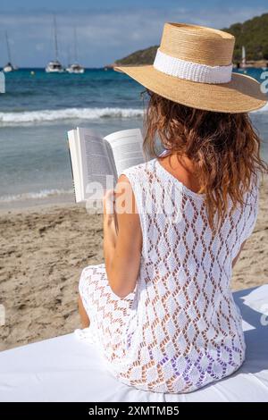 Giovane donna con abito in maglia bianca e cappello che legge sul mare sul letto bianco, Ibiza Foto Stock