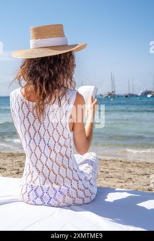 Giovane donna con abito in maglia bianca e cappello che legge sul mare sul letto bianco, Ibiza Foto Stock