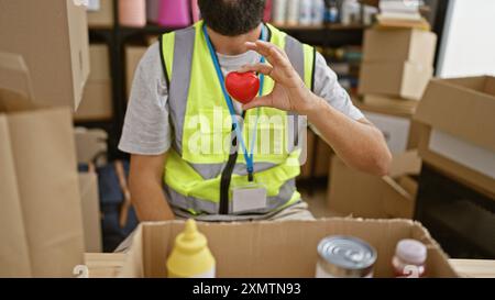 Un giovane ispanico con la barba che indossa un giubbotto di sicurezza tiene un cuore in un magazzino. Foto Stock