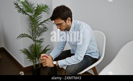 Un giovane ispanico con gli occhiali stringe una palla da stress al chiuso, seduto su una sedia in una stanza minimalista. Foto Stock