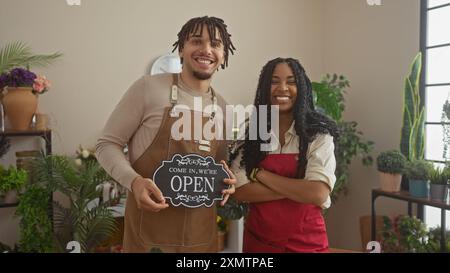 Un uomo e una donna sorridenti hanno un cartello "aperto", in piedi in un verdeggiante negozio di fiori al coperto. Foto Stock