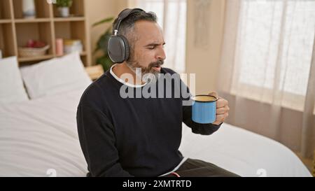 Un uomo ispanico maturo con la barba grigia si diverte ad ascoltare la musica nella sua camera da letto illuminata dal sole. Foto Stock