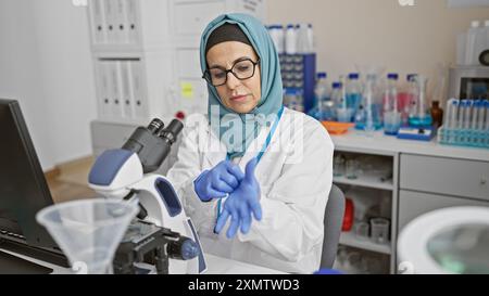 Una donna matura scienziata che indossa un hijab mette i guanti in un laboratorio con microscopi e provette. Foto Stock