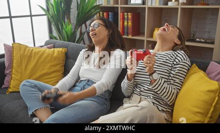 Due donne ridono gioiosamente su un divano tenendo in mano i controller di gioco in un accogliente salotto. Foto Stock