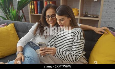 Due donne, probabilmente madre e figlia, condividono un momento guardando uno smartphone insieme, comodamente sedute in un salotto accogliente. Foto Stock