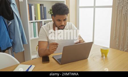 Un uomo nero concentrato esamina un documento in un ambiente ben illuminato da ufficio domestico, laptop e smartphone. Foto Stock