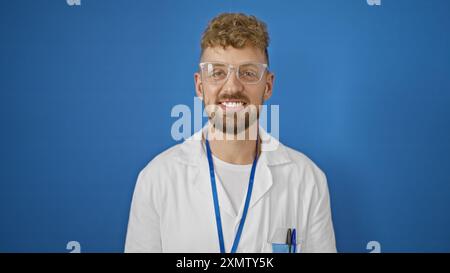 Giovane caucasico con gli occhi blu e la barba sorridente con sicurezza su uno sfondo blu isolato, indossando occhiali e un camice da laboratorio bianco. Foto Stock