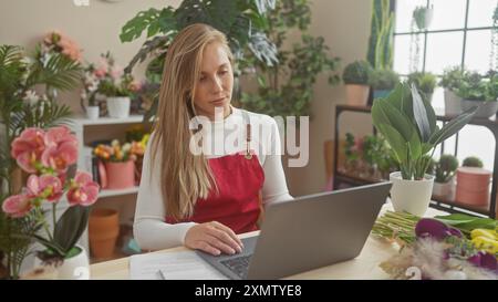 Una giovane fiorista in un negozio di fiori, circondata da piante verdi, utilizza un computer portatile in un interno colorato. Foto Stock