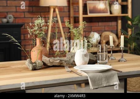Set di stoviglie pulite, vasi con bellissimi fiori, pietre e candele accese su un tavolo di legno nell'elegante sala da pranzo Foto Stock