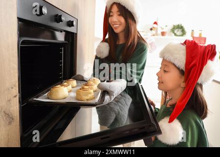 Piccola ragazza asiatica con sua madre che mette rotoli di cannella in forno in cucina la vigilia di Natale Foto Stock