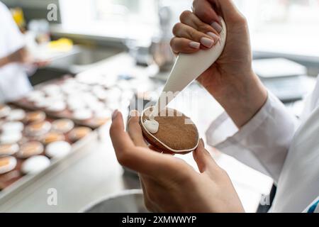 Primo piano: Mani di una pasticciera che decora i biscotti con glassa bianca mentre spreme la panna da un sacchetto. Foto di alta qualità Foto Stock