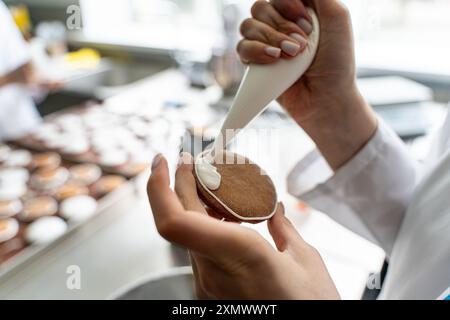Primo piano: Mani di una pasticciera che decora i biscotti con glassa bianca mentre spreme la panna da un sacchetto. Foto di alta qualità Foto Stock