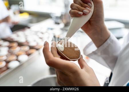 Primo piano: Mani di una pasticciera che decora i biscotti con glassa bianca mentre spreme la panna da un sacchetto. Foto di alta qualità Foto Stock